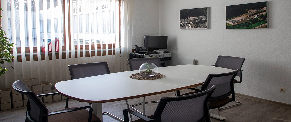 Besprechungsraum mit einem halbrunden weißen Tisch und sechs schwarzen Schwingstühlen. Auf dem Tisch steht ein durchsichiges rundes Glas mit einer kleinen grünen Pflanze, auf einem runden, braunen Untersetzer. An der weißen Wand hängen zwei Bilder die jeweils die Pappenfabrik aus älteren Jahrgängen aus der Vogelperspektive zeigen. Links im Hintergrund sind Fenster mit einem Holzrahmen, welche davor einen weißen Lamellenvorhang haben.