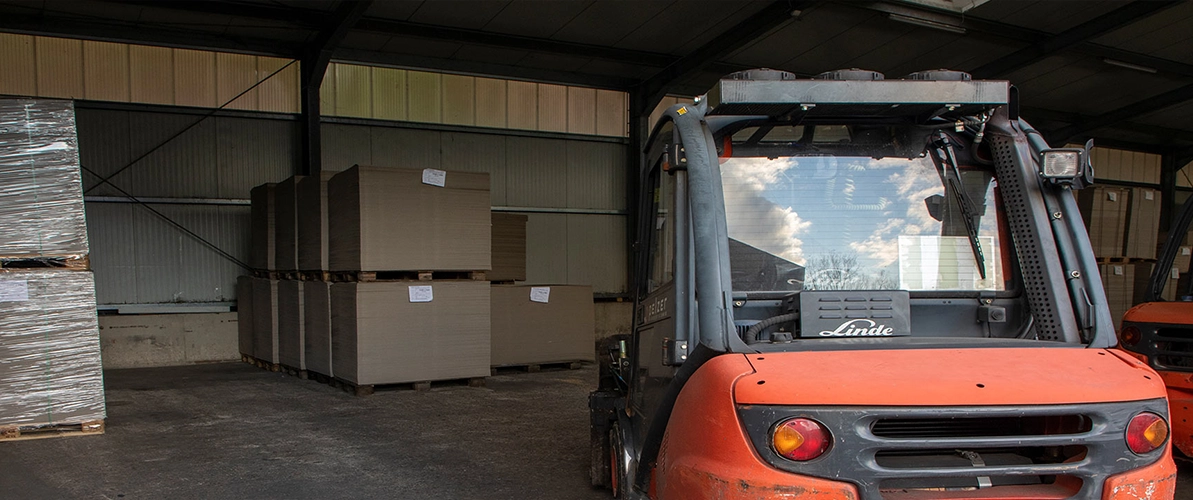 Elektro Hubstapler in schwarz-orange steht in der Lagerhalle. Im Hintergrund stehen Paletten mit Pappe beladen.