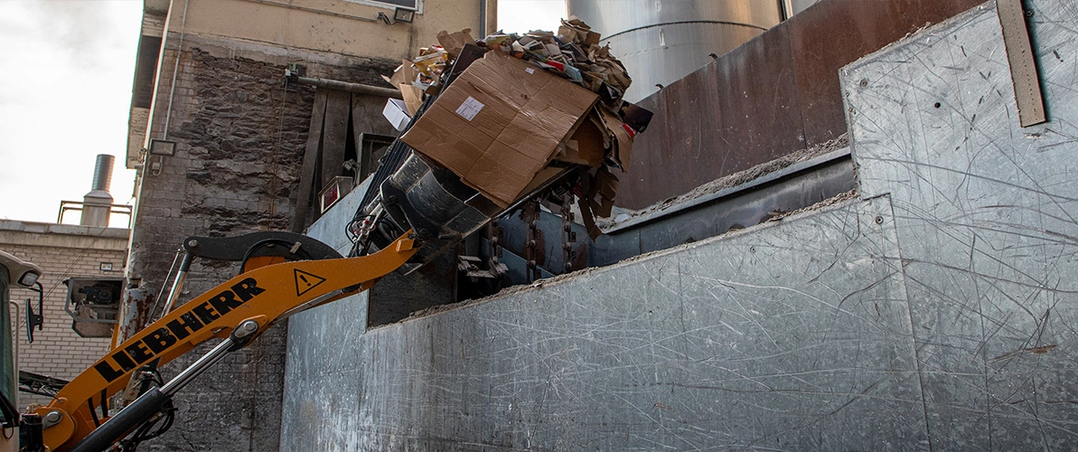Gelb-Orangefarbene Baggerschaufel, welche befüllt mit Pappresten ist, schüttet diese in eine große silberne Machine.