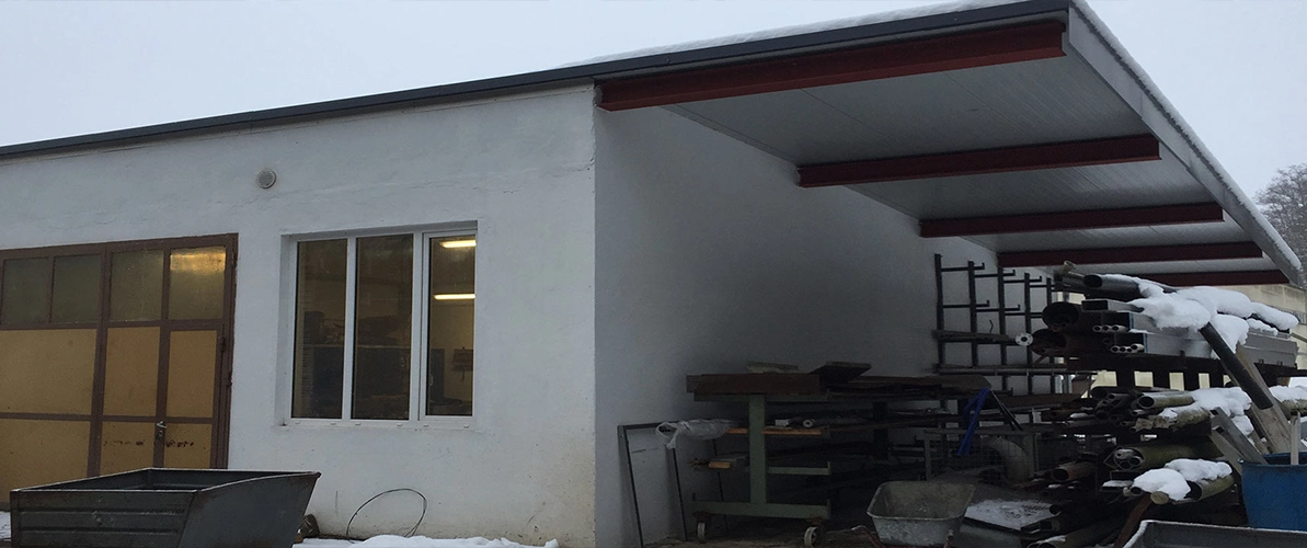 Weißes Gebäude von außen (Werkstatt) mit gelben Fesnstern. Auf der rechten Seite sind unter einer Überdachung Rohre, eine Schubkarre und weitere Dinge verstaut, welche teilweise etwas mit Schnee überzogen sind.