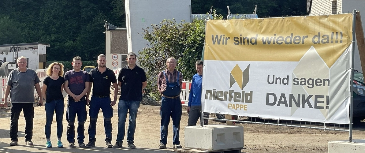 Auf der linken Seite stehen sieben Mitarbeiter:innen und rechts daneben ein gelb-weißes Schild mit der Aufschrift